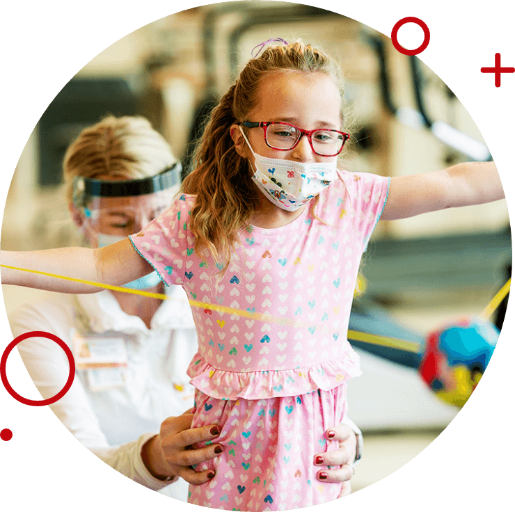 Young girl in a pink dress and face mask practicing balance with the help of a therapist in a rehabilitation setting.